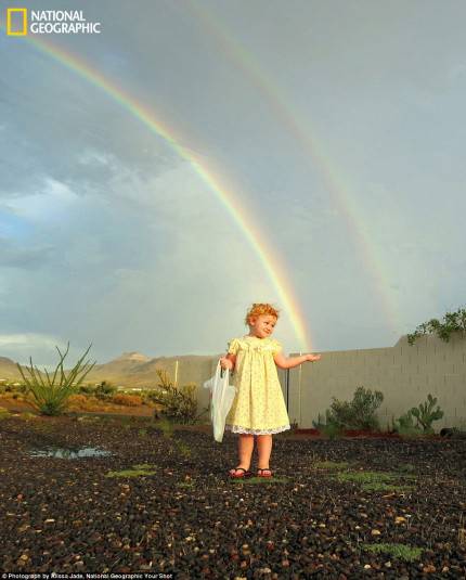 bambina di circa tre anni in abito bianco , sullo sfondo doppio arcobaleno. la mono della bambina sembra tenere un capo di arconbaleno