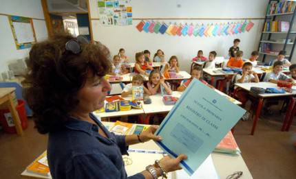 una donna insegnante in primo piano tiene registro in mano, dietro classe alunni seduti nei banchi
