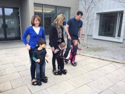 genitori che camminano insieme ai figli disabili sostenuti da una imbracatura che grazie al sostegno del loro corpo li fà camminare